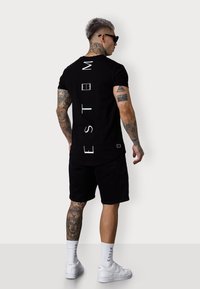 Schwarzes T-Shirt mit kurzen Ärmeln und weißem Text auf dem Rücken, kombiniert mit schwarzen Shorts und weißen Turnschuhen. Zeigt tätowierte Arme und hohe Socken.