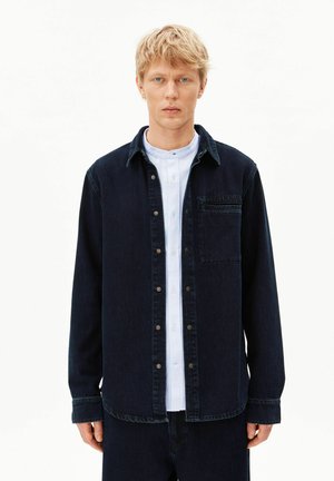 Blonder Mann mit kurzen Haaren, der ein dunkles Denim-Hemd über einem weißen Hemd mit Kragen trägt und vor einem schlichten weißen Hintergrund steht.