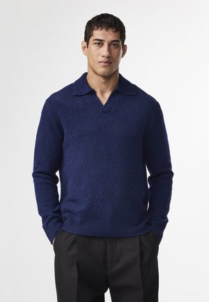 Pull en laine bleu marine avec un design col en V. Manches texturées et ajustées, et coupe décontractée au niveau des hanches. Associé à un pantalon noir.