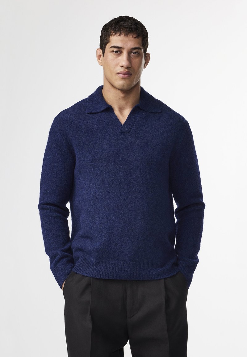 Tummansininen villapaita, jossa on kauluksellinen V-pääntie. Raskaasti teksturoitu, kapea hihansuu ja rento istuvuus lantiolla. Yhdistettynä mustiin housuihin.