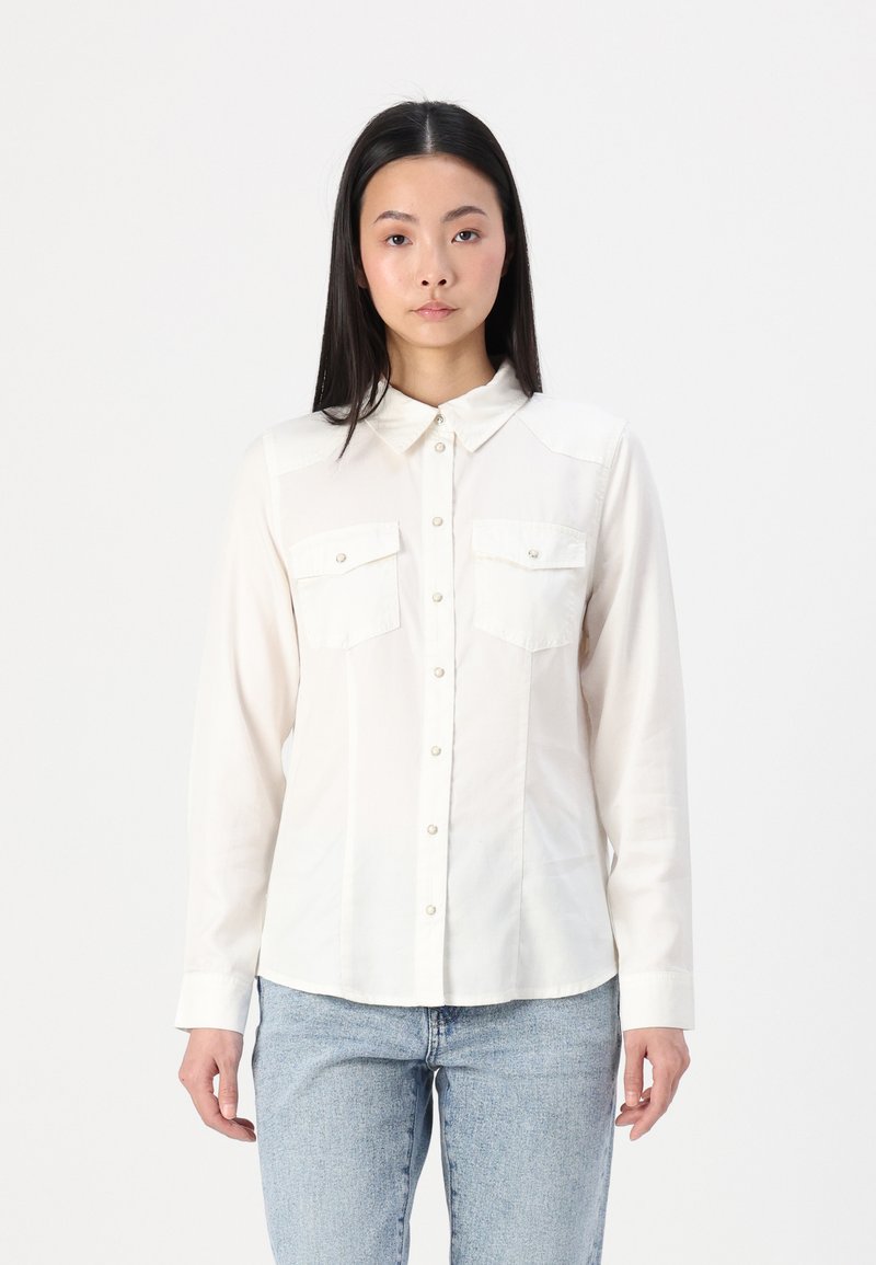Cream button-up shirt with long sleeves, two chest pockets, and a collared neck; soft fabric with a smooth texture and fitted shape.