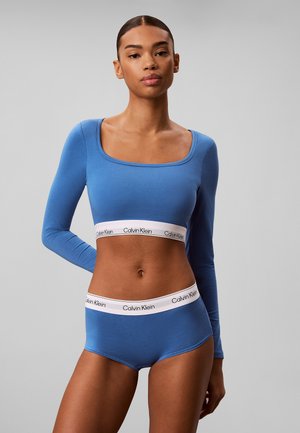 Donna che indossa un crop top blu a maniche lunghe e slip abbinati con fasce elastiche bianche con il marchio Calvin Klein.