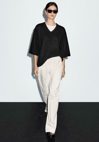 Black oversized V-neck sweater, cream pants, and black shoes; model also wears rectangular sunglasses. Smooth texture, relaxed fit.