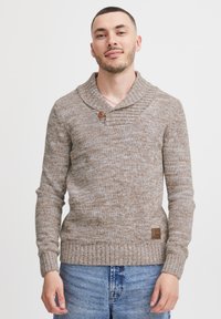Brun strikket sweater med sjalskrave, der har en træknap, ribbede manchetter og et struktureret mønster. Kombineret med blå denim jeans.