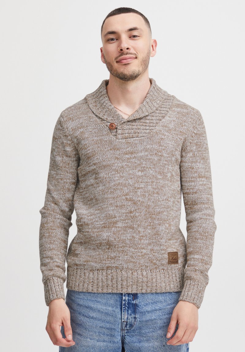 Brązowy sweter dzianinowy z kołnierzem szalowym, z drewnianym guzikiem, ściągaczami na rękawach oraz teksturowanym wzorem. Połączony z niebieskimi dżinsami.