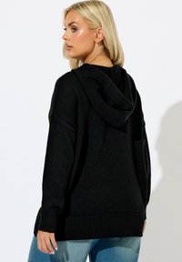 Felpa nera in maglia con una vestibilità rilassata, orlo a costine e un grande cappuccio. La texture appare morbida, ideale per essere indossata sopra abbigliamento casual.