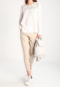 Ljusbeige långärmad topp med spetsmönster vid halsringningen, tillsammans med beige slim-fit byxor och vita sneakers. Håller en ljusgrå ryggsäck.