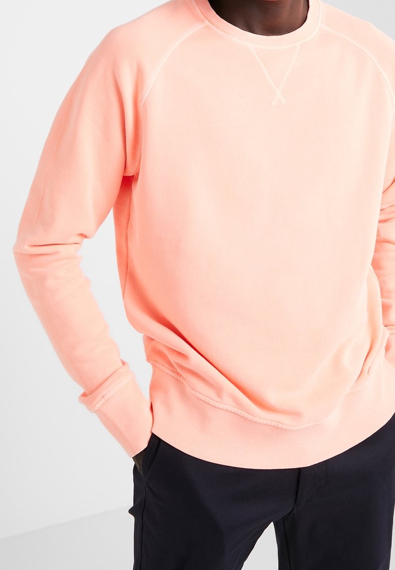 Personne portant un sweat-shirt à manches longues rose clair, les mains dans les poches d’un pantalon foncé, debout devant un fond uni.
