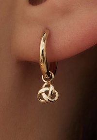 Gold hoop earring with a pendant featuring an intricate knot design; smooth, shiny surface; mid-sized hoop; hangs near the earlobe.