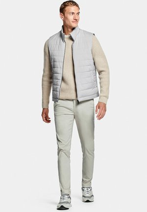 Man loopt, draagt een lichtgrijze doorgestikte bodywarmer, een beige trui, lichtgrijze broek en grijze sneakers, kijkt naar de zijkant en lacht.