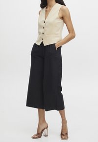 Beige bezrukávový svetr s výstřihem do V, pěti černými knoflíky a dvěma předními kapsami, zkombinovaný s volnými černými kalhotami širokého střihu a průhlednými sandály na blokovém podpatku.