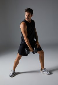 Black sleeveless athletic shirt and shorts, gripping gray dumbbells, against a neutral gray background. Athletic footwear with reflective accents.