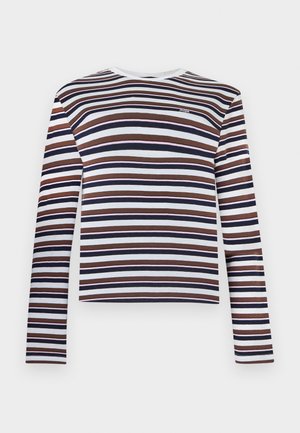 Kobieta ubrana w pasiastą bluzkę z długim rękawem, beżowe szorty, białe skarpetki, brązowe buty na platformie oraz niosąca czarną torbę na ramię z frędzlami.