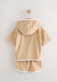 Set color cammello in spugna, composto da una maglia con cappuccio a maniche corte e pantaloncini abbinati, bordati con rifiniture bianche. Tessuto liscio, design casual.