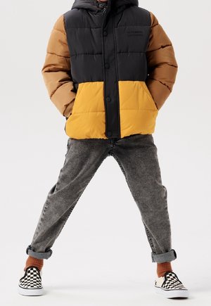 Puffjacke mit einem Farbblockdesign in Schwarz, Braun und Gelb, ausgestattet mit Kapuze, Druckknöpfen und gestepptem Material. Kombiniert mit grauen Jeans und karierten Slippern.