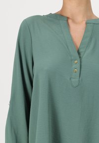 Camicia verde leggera con scollo a V, tre bottoni dorati e maniche lunghe. Il tessuto ha una texture liscia e una leggera caduta.