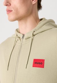 Felpa verde chiaro con zip, caratterizzata da una texture morbida, cappuccio con cordoncino e un'etichetta logo rettangolare rossa con testo nero.