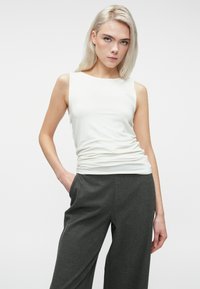 Femme blonde aux cheveux raides portant un haut sans manches blanc et un pantalon ample gris foncé, se tenant avec une main dans la poche.