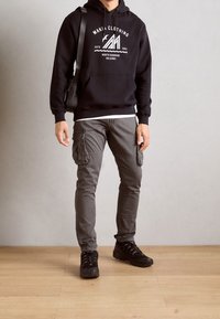 Czarna bluza z kapturem z białym logo graficznym, zestawiona z szarymi spodniami cargo i czarnymi butami na sznurówki. Casualowy strój na świeżym powietrzu.