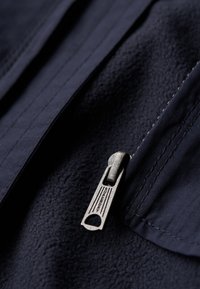 Marineblå fleece-stof med en glat tekstur; har en metallisk lynlås med "Superdry" branding og syningsdetaljer.
