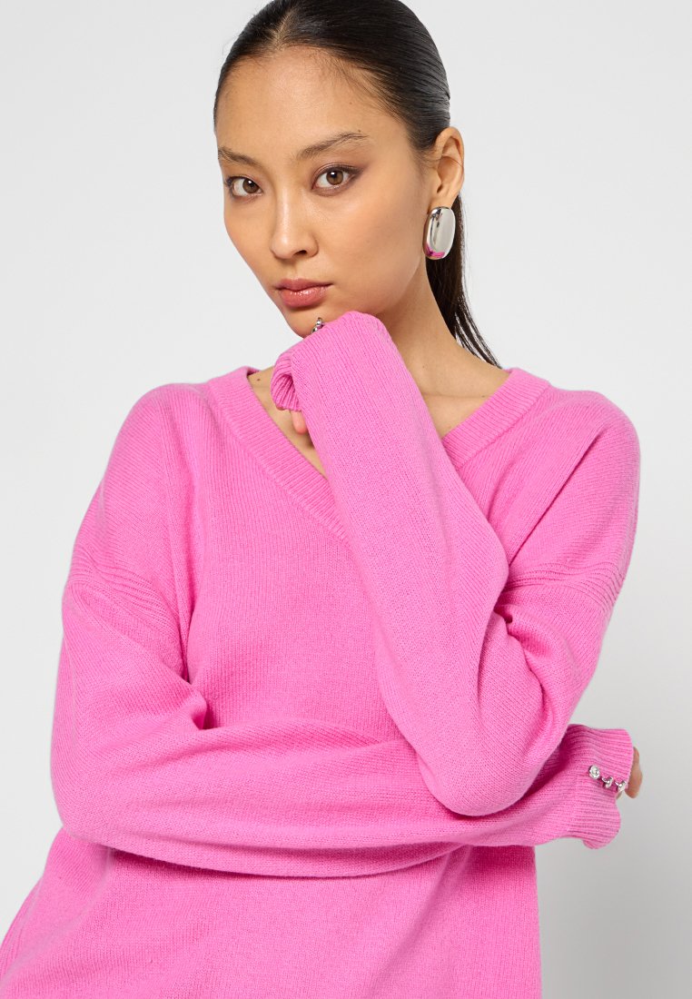Mujer con cabello oscuro, vestida con un suéter de pico rosa, que lleva pendientes ovalados de plata, posando con la mano cerca de su cara sobre un fondo sencillo.