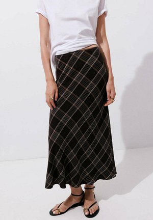 Mujer con camiseta blanca de manga corta, falda midi de cuadros en negro y gris, anillos dorados y sandalias negras con tira en el tobillo, de pie sobre suelo blanco.
