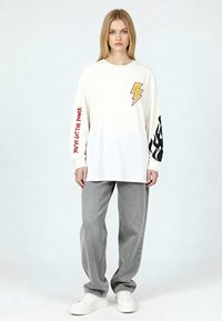 Femme blonde portant une chemise blanche à manches longues avec un éclair jaune, un texte rouge "Tu as le pouvoir", un motif de flammes noires, un jean gris et des baskets blanches.