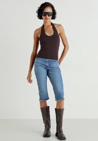Top halter maro, blugi scurți bleu deschis și cizme maro până la genunchi. Ochelari de soare negri, eleganți. Texturi fine și siluetă casual.