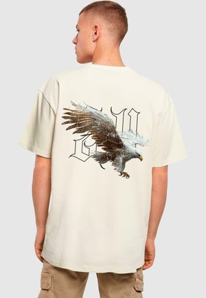 Beige bomulds t-shirt med et stort ørner grafik på ryggen, med detaljerede udsmykninger og fedt sort skrift over designet.