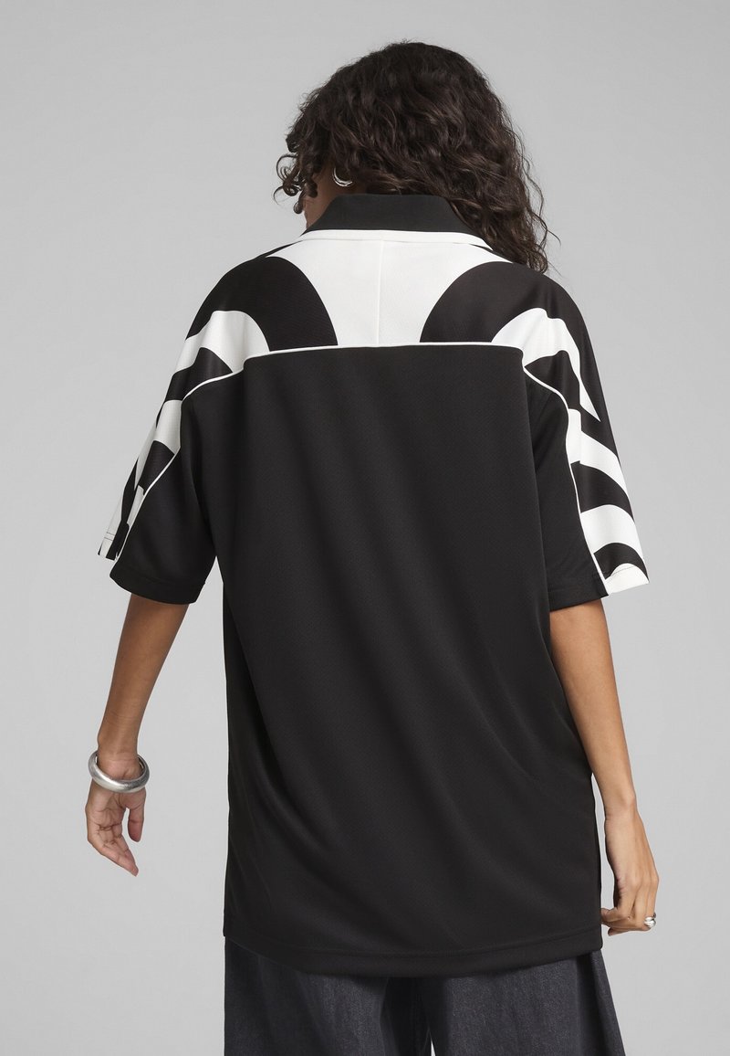 Personne aux cheveux bouclés porte une chemise noire oversize avec des motifs graphiques blancs sur les manches et les épaules, vue de dos.