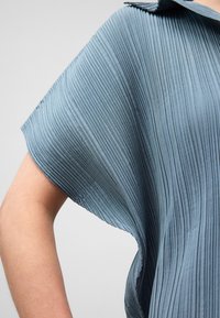 Blusa plissettata in morbido tessuto blu, con ampio colletto e vestibilità ampia, caratterizzata da una texture verticale e una leggera lucentezza.