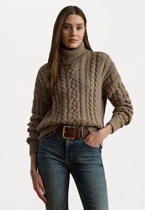 Chunky beige knit turtleneck sweater with cable patterns, paired with blue jeans and a brown belt, showing a relaxed fit and textured design.