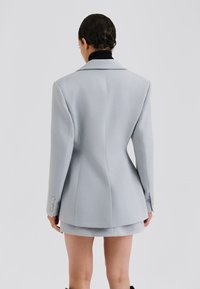 Blazer gris clair cintré avec un ajustement slim, de larges revers et des boutons dorés aux manches, présentant un tissu lisse et texturé.