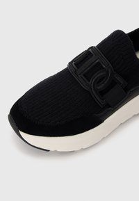 Svart sneaker med teksturert stoff og semsket skinn-detaljer, som har en bred svart spenne og en tykk hvit såle.