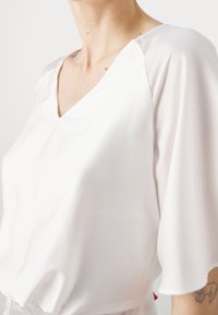 Blouse en soie blanche avec un col en V et des manches courtes douces et fluides. Le tissu a une texture lisse, reflétant la lumière de manière subtile.