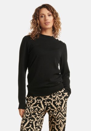 Black long-sleeve sweater with a high neckline, ribbed cuffs, and decorative shoulder buttons, paired with patterned black and cream trousers.