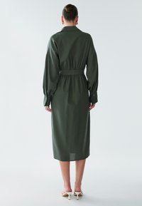 Žalia ilgomis rankovėmis suknelė su diržu juosmens srityje, saga užsegamomis rankogaliais ir apykakle. Audinys yra lygios tekstūros, o kirpimas tiesus, siekiantis vidurinį blauzdos aukštį.