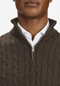 Brun kabelstickad tröja med dragkedja i kragen, bärs över en vit skjorta med krage. Har ett upphöjt, strukturerat mönster och silverfärgad dragkedja.