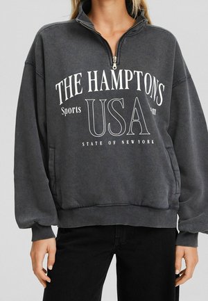 Ciemnoszara bluza z półzamkiem, z nadrukiem "THE HAMPTONS USA State of New York" w białym kolorze, noszona z czarnymi spodniami.