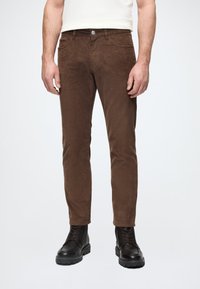 Καφέ παντελόνι κόρντουρα με στενή γραμμή, υφασμάτινη υφή, μπροστινές τσέπες και κλείσιμο με μεταλλικό κουμπί, συνδυασμένο με μαύρες μπότες με κορδόνια.