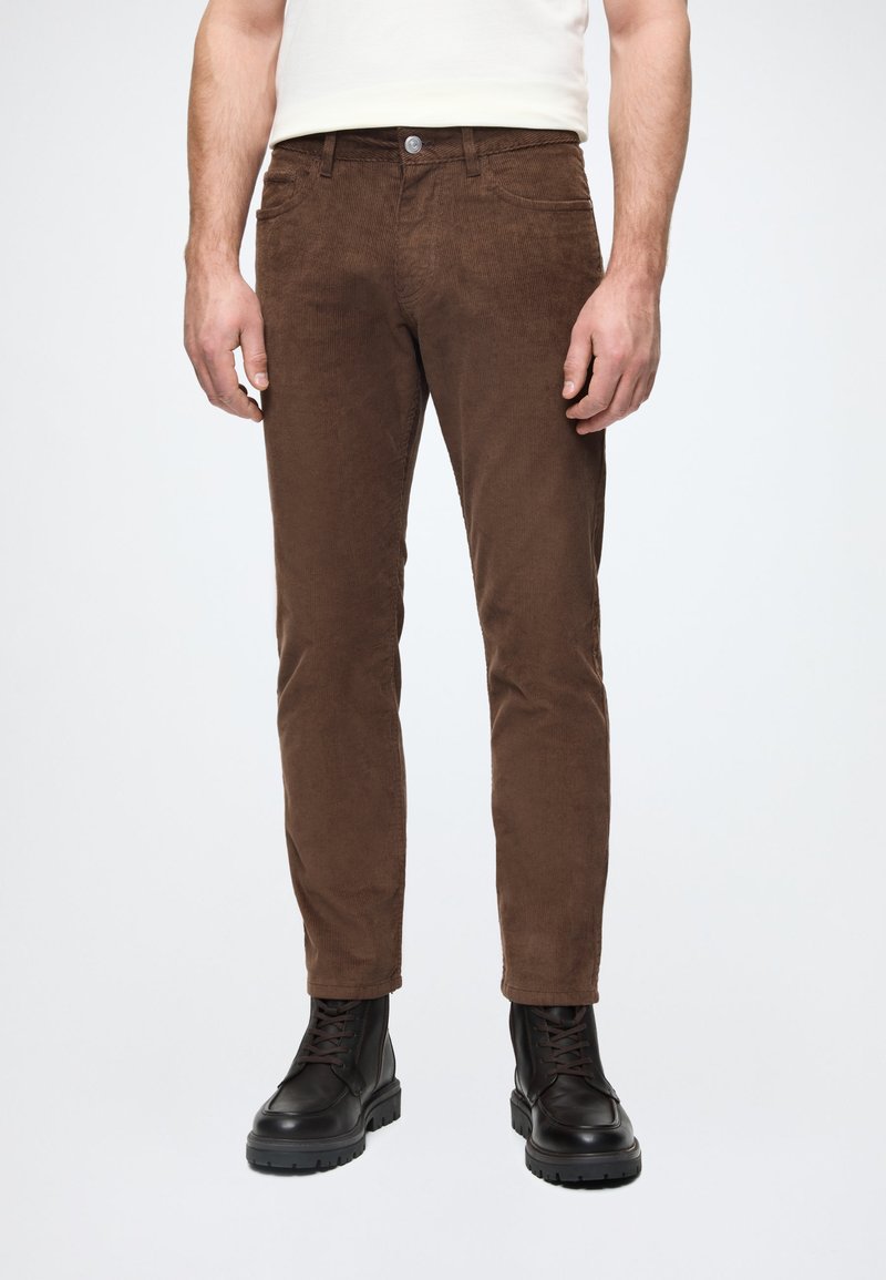 Καφέ παντελόνι κόρντουρα με στενή γραμμή, υφασμάτινη υφή, μπροστινές τσέπες και κλείσιμο με μεταλλικό κουμπί, συνδυασμένο με μαύρες μπότες με κορδόνια.
