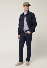 Chaqueta de algodón navy sobre una camisa de rayas azul claro, combinada con pantalones navy y mocasines grises. Gorra oliva llevada encima.