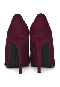 Un paio di scarpe col tacco bordeaux con punte affusolate, viste da dietro su uno sfondo bianco.