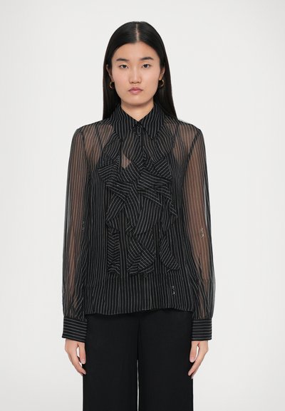 Blusa negra transparente con rayas verticales, que presenta un delantero con volantes, cierre de botones y mangas largas con acentos en los puños. Combinada con pantalones negros.