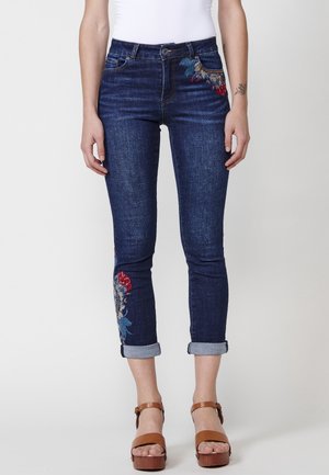 Frau trägt dunkelblaue hochgekrempelte Jeans mit Blumenstickerei am Oberschenkel und an der Wade, kombiniert mit braunen Plateausandalen.