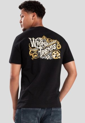T-shirt en coton noir avec un graphisme coloré "MAUVAISES AMIES" au dos, avec des détails ornés en blanc et or. Manches courtes.