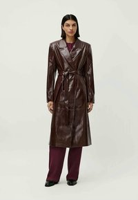 Casaco longo de pele castanha com cintura com cinto, lapelas recortadas e acabamento brilhante, combinado com calças escuras de cor burgundy e sapatos.