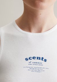 Witte geribde tanktop met een ronde hals; heeft blauwe tekst "geuren van de zomer" op de voorkant, wat een textuur detail creëert.
