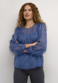 Blå texturerad blus med genomskinligt tyg och fransdesign, långa ärmar och rundad halsringning, kombinerad med mörkgrå jeans.