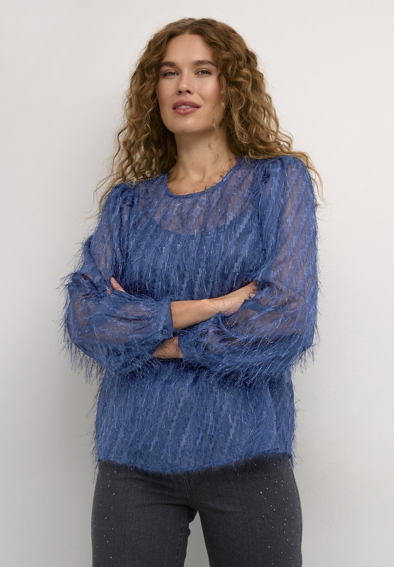 Blå texturerad blus med genomskinligt tyg och fransdesign, långa ärmar och rundad halsringning, kombinerad med mörkgrå jeans.